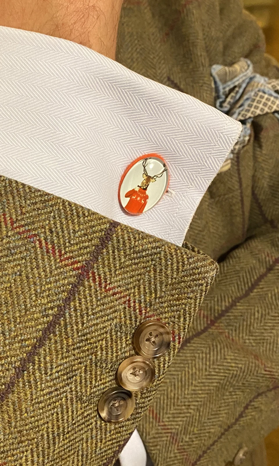 Ovaler Manschettenknopf mit Hirsch im roten Pullover an einer weißen Manschette unter einem Tweedsakko