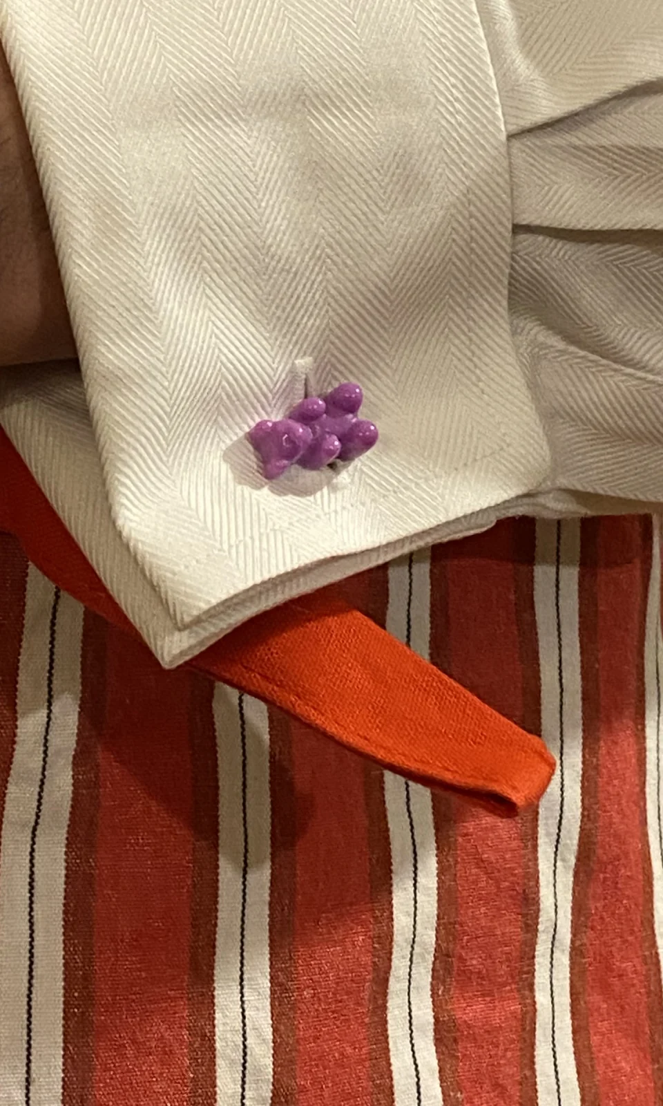 Manschettenknopf Manschettenbär in Violett an einer weißen Manschette vor rot gestreiftem Stoff